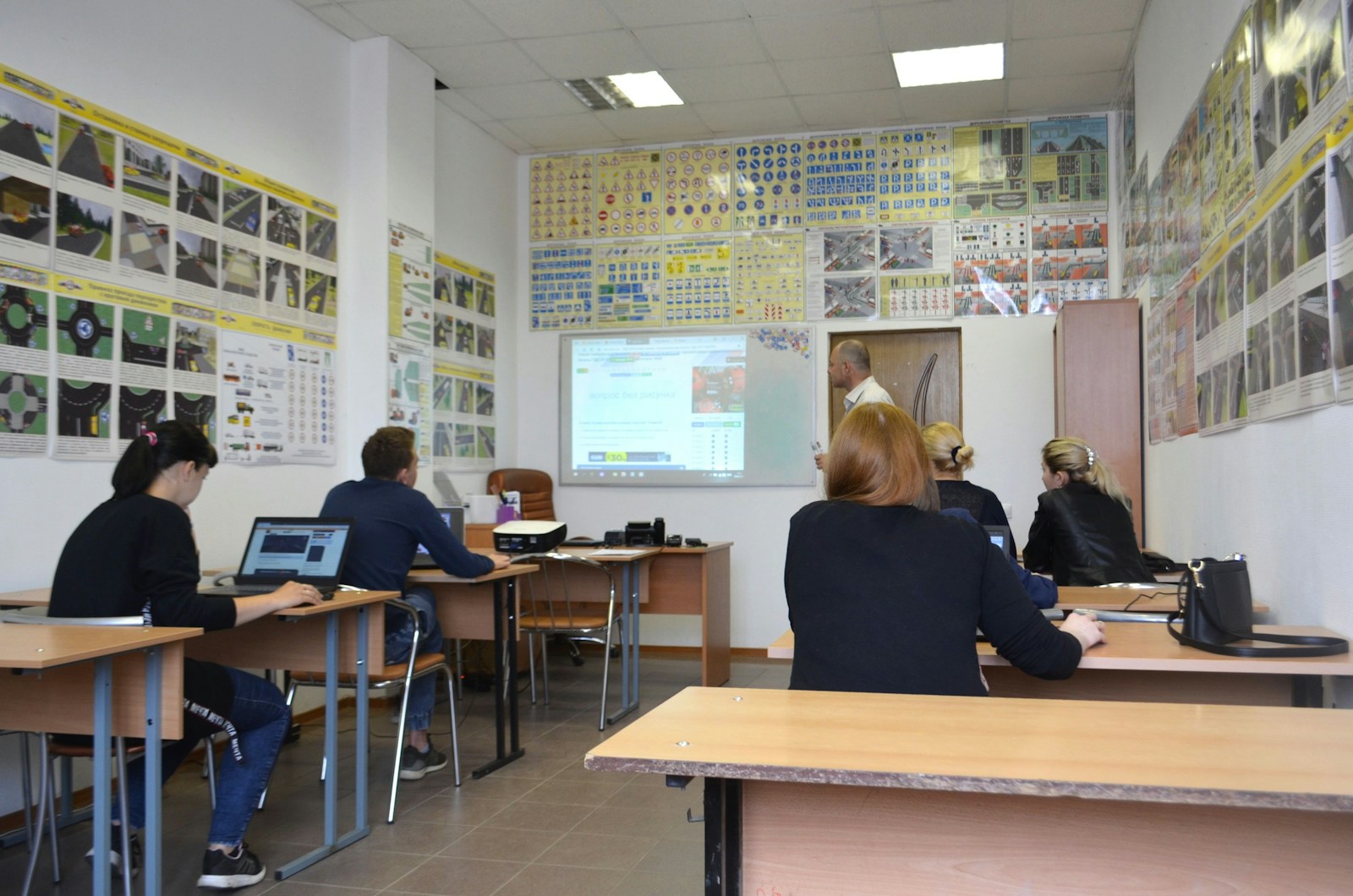 Salle de cours vue de l'arrière, plusieurs personnes assises devant des ordinateurs portables suivent une présentation projetée au mur